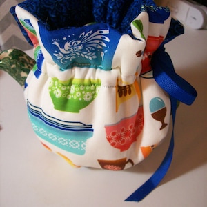 Puede incluir: Una funda para tetera blanca con una cinta azul. La funda está decorada con un patrón colorido de tazas de té y un pavo real.