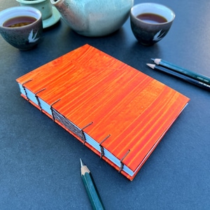 Könnte beinhalten: Ein leuchtend orangefarbenes Notizbuch mit Holzmaserung und braunen Nähten. Das Notizbuch liegt geschlossen auf einer dunkelgrauen Oberfläche mit zwei Bleistiften daneben.