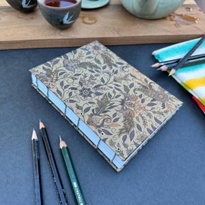 May include: A brown and beige journal with a floral pattern on the cover. The journal is closed and lying on a dark gray surface. There are three pencils lying next to the journal.