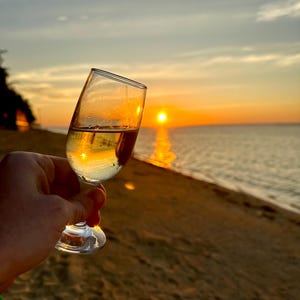 Puede incluir: Una copa de vino transparente, llena de un l&iacute;quido claro, se sostiene frente a una puesta de sol sobre el mar. El cristal refleja los tonos dorados del sol poniente, creando una escena c&aacute;lida. La playa y el cielo son visibles en el fondo.