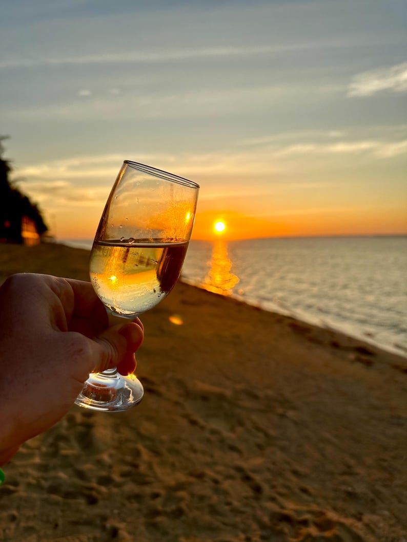 Puede incluir: Una copa de vino transparente, llena de un l&iacute;quido claro, se sostiene frente a una puesta de sol sobre el mar. El cristal refleja los tonos dorados del sol poniente, creando una escena c&aacute;lida. La playa y el cielo son visibles en el fondo.