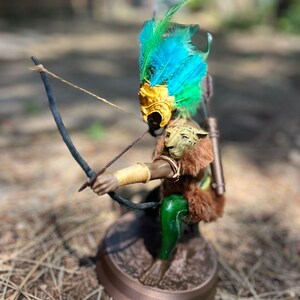 以下が含まれることがあります： 羽根飾りをかぶった神話上の生き物の置物で、弓矢を持っています。生き物は金色のマスク、緑と青の羽根、茶色の毛皮の体を持っています。置物は茶色の台座の上に立っています。