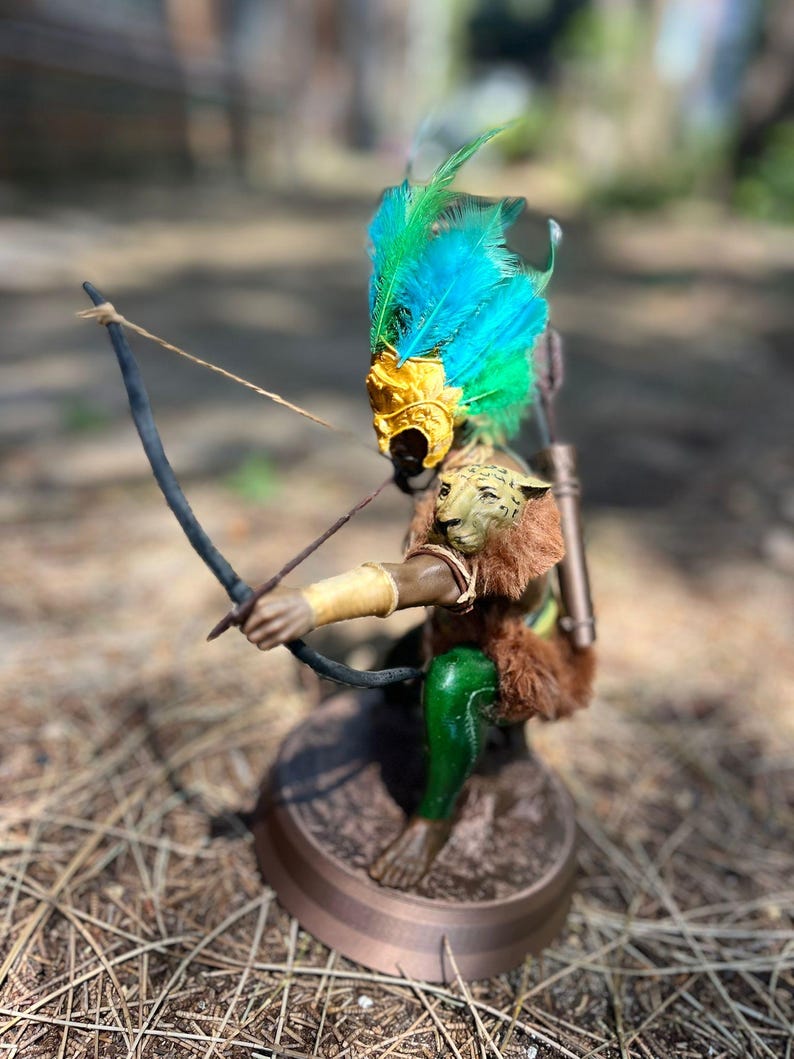 以下が含まれることがあります： 羽根飾りをかぶった神話上の生き物の置物で、弓矢を持っています。生き物は金色のマスク、緑と青の羽根、茶色の毛皮の体を持っています。置物は茶色の台座の上に立っています。