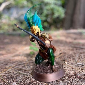以下が含まれることがあります： 羽根飾りをかぶった緑と茶色の架空の生き物の置物で、弓矢を持っています。置物は茶色の台座の上に立っています。