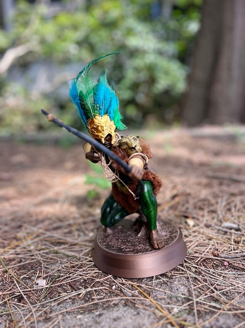 以下が含まれることがあります： 羽根飾りをかぶった緑と茶色の架空の生き物の置物で、弓矢を持っています。置物は茶色の台座の上に立っています。
