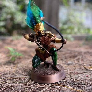 以下が含まれることがあります： 羽根飾りをかぶった人物の緑と茶色の置物で、弓矢を持っています。置物は茶色の台座の上に立っています。