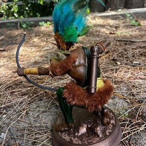 以下が含まれることがあります： 弓矢を持った人物の茶色と緑の置物。人物は羽根飾りをかぶり、毛皮の縁取りのある衣服を着ている。置物は茶色の台座の上に立っている。