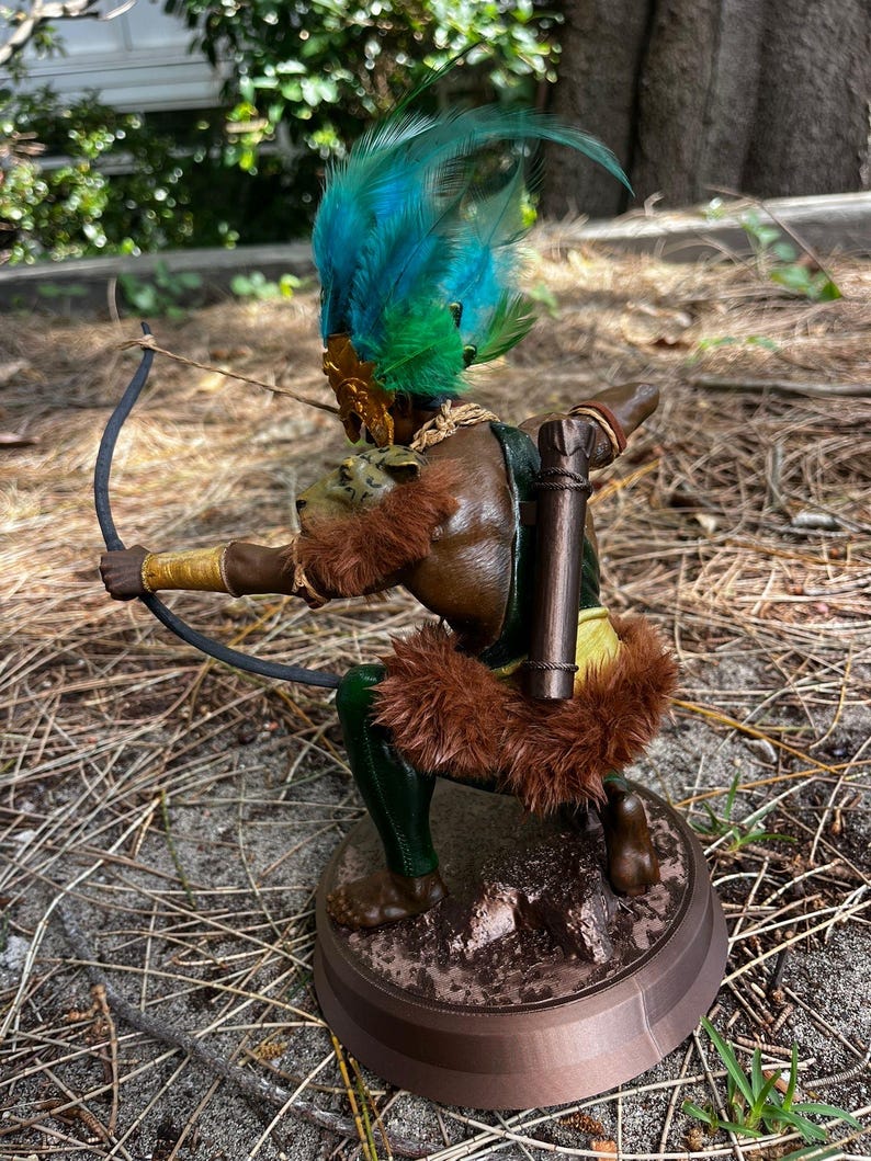 以下が含まれることがあります： 弓矢を持った人物の茶色と緑の置物。人物は羽根飾りをかぶり、毛皮の縁取りのある衣服を着ている。置物は茶色の台座の上に立っている。