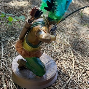 以下が含まれることがあります： 羽根飾りをかぶったひざまずく弓矢を持った弓射手の置物。弓矢は茶色のチュニック、緑色のズボン、金色のベルトを身につけている。弓矢は右手に弓と矢を持っている。置物は茶色の台座の上に置かれている。