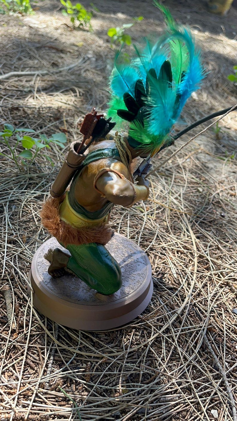 以下が含まれることがあります： 羽根飾りをかぶったひざまずく弓矢を持った弓射手の置物。弓矢は茶色のチュニック、緑色のズボン、金色のベルトを身につけている。弓矢は右手に弓と矢を持っている。置物は茶色の台座の上に置かれている。