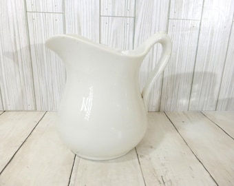 May include: A solid off-white ceramic pitcher with a smooth, glossy finish. The vessel features a rounded, bulbous body, a wide pouring spout, and a curved handle attached to the side. The design is simple, functional, and monochromatic.