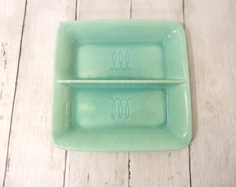 May include: A square, light mint green ceramic divided dish with rounded corners. The dish features a central horizontal divider and two identical embossed cursive letter M designs, one centered in each of the two rectangular compartments.