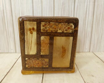 May include: A rectangular resin block with a dark brown frame containing embedded dried botanical elements. The design features sections with small round seeds, thin stems with flower buds, a textured floral piece, and a single leaf.
