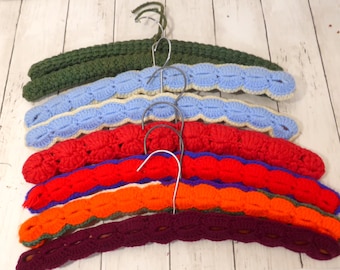 May include: A set of six clothes hangers, each covered in a crocheted yarn sleeve. The hangers feature a variety of solid colors including dark green, light blue, bright red, orange, and deep purple, all equipped with silver metal hooks.