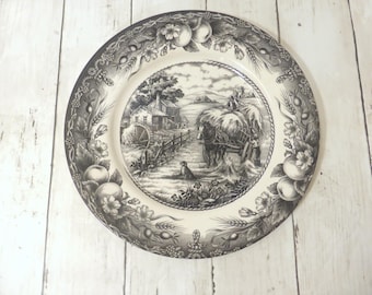 May include: A round ceramic plate featuring a black and white transferware scene of a horse-drawn hay wagon, a water mill, and a dog. The rim is decorated with a detailed border of apples, wheat stalks, and flowers in a matching monochrome style.