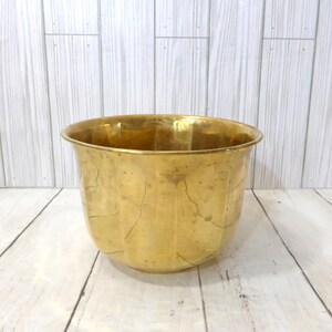 May include: A polished brass planter with a wide, circular opening and a rounded body featuring vertical fluted panels. The metal surface displays a warm, golden tone with subtle variations in texture and natural signs of aging throughout.