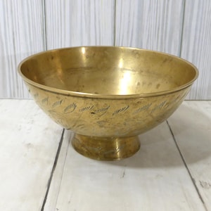 May include: A polished brass bowl with a wide, circular opening and a pedestal base. The exterior surface of the bowl features a series of etched, repeating leaf-like patterns that encircle the body, showing a warm, metallic gold tone.