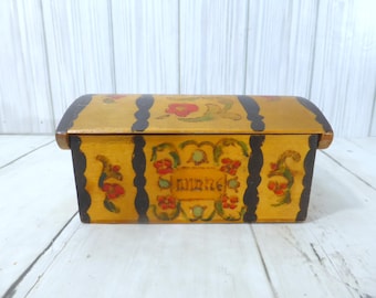 May include: A rectangular wooden box with a curved lid, painted in a mustard yellow finish. The box features black vertical stripes and hand-painted floral motifs in red, green, and blue. The word "minne" is inscribed in the center front.