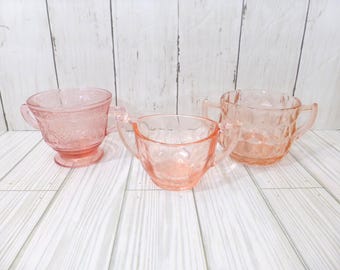 May include: Three pink depression glass sugar bowls with two handles each. The bowls feature distinct pressed patterns including floral motifs, scalloped edges, and geometric diamond textures on the exterior surfaces of the translucent pink glass vessels.