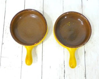 May include: Two round ceramic bowls with handles, featuring a bright yellow exterior and a smooth, dark brown glazed interior. The bowls are positioned side by side, showcasing their circular shape and the contrast between the yellow and brown.