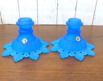 May include: A pair of bright blue frosted glass candle holders featuring a wide, scalloped base with intricate leaf-like cutouts and a smooth, cylindrical top section. Each piece displays a small, circular gold-toned sticker on the front.