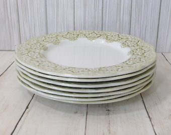 May include: A stack of five round white ceramic dinner plates featuring a wide rim decorated with an intricate olive green floral damask pattern. The plates have a smooth, glossy finish and a scalloped edge design, creating a uniform appearance.