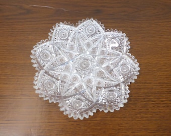 May include: A clear, circular glass dish featuring an intricate, multi-pointed star pattern with deep, geometric cross-hatch cuts and a serrated, scalloped edge. The design radiates from the center, creating a complex, faceted light-reflecting surface.