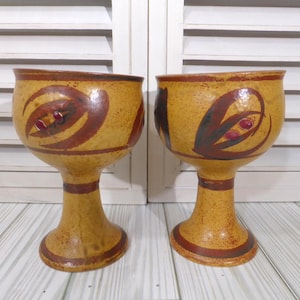 May include: Two ceramic goblets featuring a speckled mustard yellow glaze with hand-painted dark brown abstract leaf motifs and small red accents. Each goblet has a wide bowl, a tapered stem, and a circular base with brown painted bands.