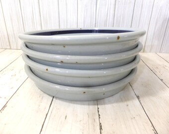 May include: A stack of four round, shallow enamelware plates with a light gray exterior and a dark blue interior rim. The plates feature a smooth, glossy finish with visible small brown spots of wear along the outer edges of the rims.