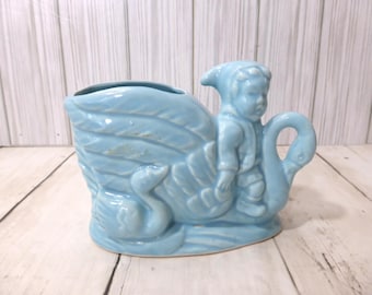 May include: A light blue ceramic planter shaped like a swan boat with a figure wearing a hood seated inside. The vessel features molded feather details on the sides and a smaller swan head at the front, finished with a glossy, smooth glaze.