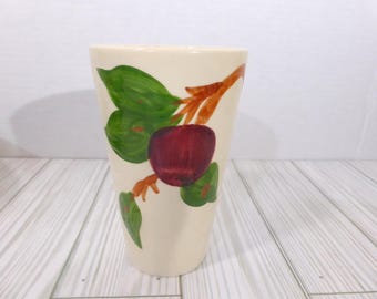 May include: A cream-colored ceramic vase featuring a hand-painted design of a dark red apple on a brown branch with vibrant green leaves. The vase has a tapered, conical shape with a smooth finish and a wide, open top rim for floral arrangements.