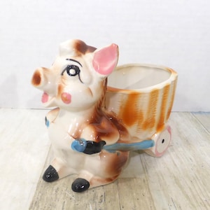 Könnte beinhalten: Ein Keramik-Blumentopf in Form eines Schweins, das einen gestreiften Wagen zieht. Das Schwein ist cremefarben mit braunen und rosa Akzenten, schwarzen Hufen und blauen Griffen. Der Wagen ist orange und weiß mit rosa Rädern.