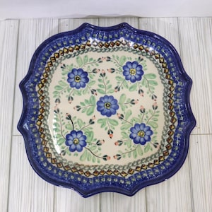 May include: A ceramic serving platter with a scalloped edge and a floral design. The platter features a blue border with a detailed pattern, surrounding a cream-colored center with blue flowers and green leaves.