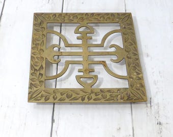May include: A square brass trivet featuring an intricate openwork design in the center and a decorative border embossed with a repeating leaf pattern. The metal surface displays a warm, metallic gold tone with subtle signs of natural aging.