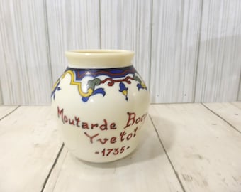 May include: A cream-colored ceramic mustard jar with a rounded body and a wide opening. The jar features a decorative blue, red, and yellow floral border around the neck and the text "Moutarde Boco Yvetot -1735-" printed in dark red lettering.