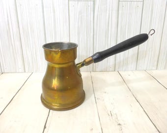 May include: A brass coffee pot with a wide base, a narrow neck, and a flared rim. The pot features a long, black wooden handle attached with a brass-colored metal ferrule. A small metal loop is located at the end of the handle for hanging.