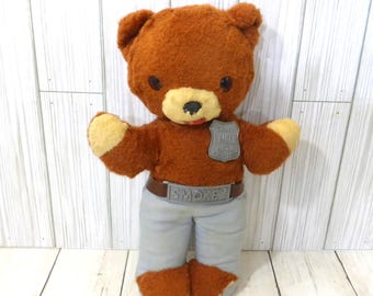 May include: A brown plush bear toy with a cream-colored muzzle and paw pads. The bear wears light gray pants, a brown belt with a silver buckle labeled SMOKEY, and a silver shield-shaped badge on its chest with the text SMOKEY BEAR.