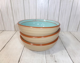 May include: A stack of three round ceramic bowls with a tan exterior and a smooth, light turquoise interior. Each bowl features a dark orange rim and a small, decorative geometric pattern in white and dark orange along the inner edge.