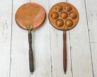 May include: Two copper pans with long wooden handles. The left pan has a circular, hammered surface. The right pan features seven circular indentations for cooking, arranged in a hexagonal pattern. Both pans have brass hardware connecting the handles.