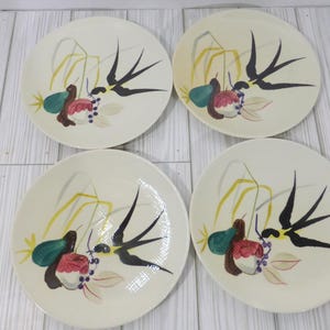 Peut inclure: Un ensemble de quatre assiettes rondes en céramique blanc cassé, chacune ornée d'un motif peint à la main représentant une hirondelle noire en vol, des tiges de blé jaunes, une poire verte, une pomme rouge et des petites baies violettes.