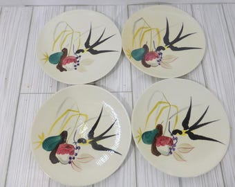May include: A set of four round off-white ceramic plates, each featuring a hand-painted design of a black swallow in flight, yellow wheat stalks, a green pear, a red apple, and small purple berries arranged in a cluster on the surface.