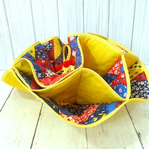 May include: A fabric organizer with multiple compartments featuring a bright yellow interior and a patchwork exterior pattern of red, blue, and yellow floral and geometric prints. The edges are finished with yellow binding and a small red accent.