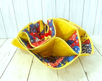 May include: A fabric organizer with multiple compartments featuring a bright yellow interior and a patchwork exterior pattern of red, blue, and yellow floral and geometric prints. The edges are finished with yellow binding and a small red accent.