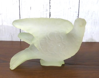 May include: A pale yellow, translucent glass figurine shaped like a bird in flight. The object features a frosted, matte texture with molded details depicting feathers on the wings and tail. The bird is captured in a side profile view with its wings extended upward and its tail feathers pointing downward.