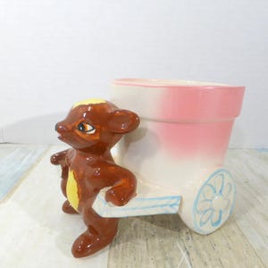 Puede incluir: Una maceta de cerámica vintage con una figura de oso marrón tirando de una maceta en forma de carro. La maceta es blanca con un efecto degradado rosa, y las ruedas tienen un diseño floral azul. Una pieza decorativa encantadora.