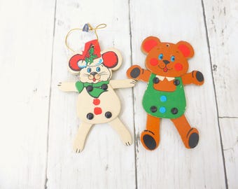 May include: Two flat wooden ornaments featuring a cream-colored mouse wearing a green scarf and a red hat with holly, and a brown bear wearing green overalls with blue buttons. Both ornaments have articulated limbs and a gold hanging string.