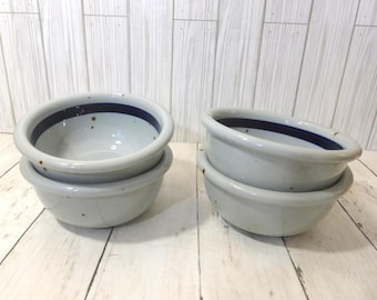 May include: Four ceramic bowls arranged in two stacks of two. Each bowl features a light gray speckled glaze with a prominent dark blue band circling the interior rim. The bowls have a rounded, thick-walled design with a smooth, glossy finish.