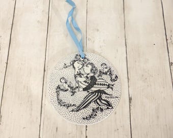 May include: A circular white ceramic ornament featuring a detailed black ink illustration of two figures embracing, surrounded by small floral patterns and a cherub. A light blue ribbon is attached to the top for hanging purposes.