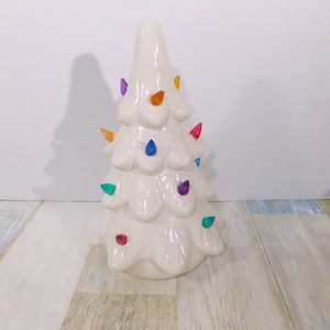 May include: A ceramic Christmas tree in a glossy off-white finish. The tree is adorned with colorful, teardrop-shaped lights in red, orange, yellow, green, blue, and purple. The tree has a classic, tiered design, perfect for holiday decor.
