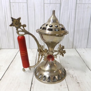May include: A brass incense burner featuring a metallic finish, a dome-shaped lid with circular cutouts, and a decorative handle with a cylindrical red grip. The base is flared with triangular cutouts, and the structure includes leaf-shaped accents.